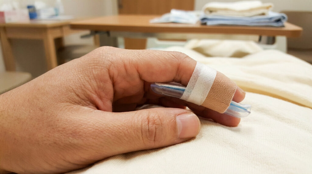 Main avec doigt immobilisé par attelle et pansement, posée sur un drap. Arrière-plan flou d'une chambre d'hôpital.