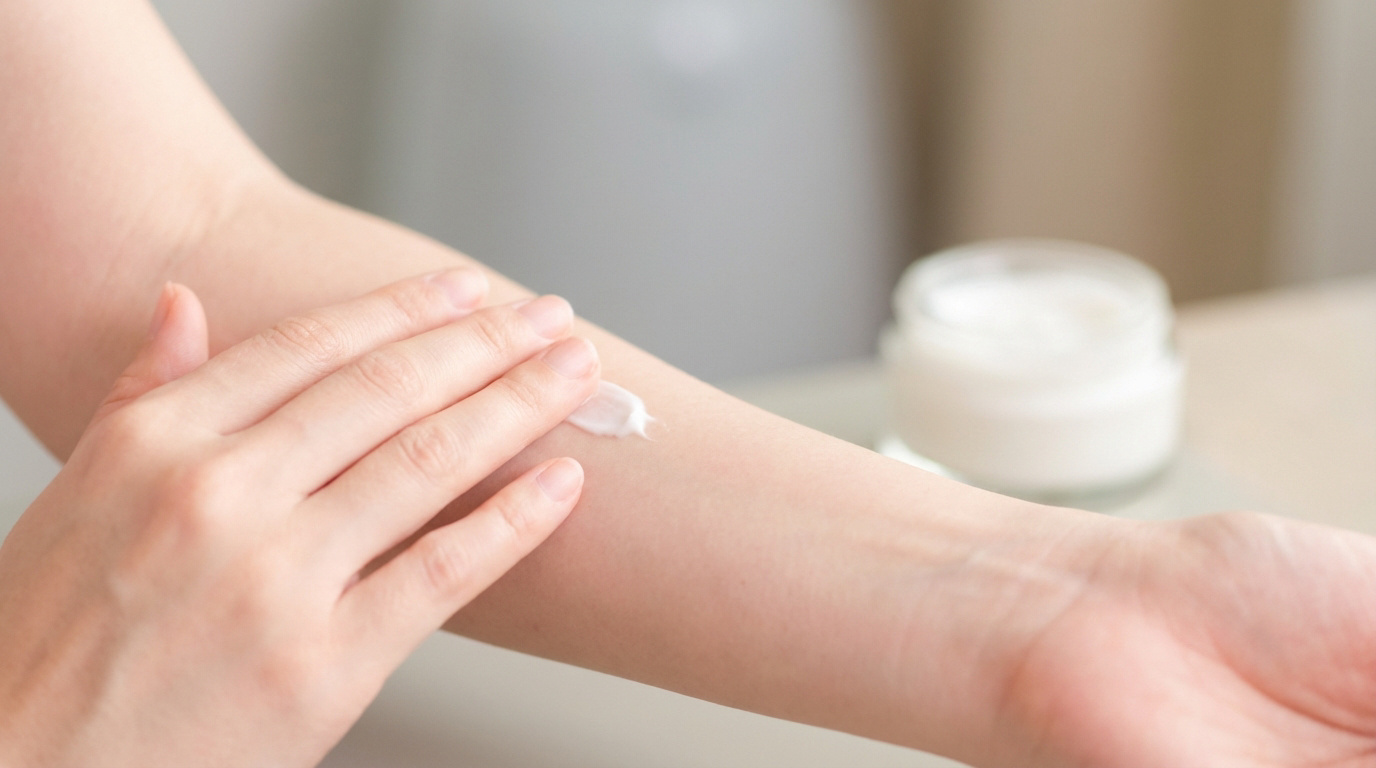 Une main applique de la crème blanche sur un bras, un pot de crème est visible en arrière-plan flou. Image illustrant le soin d'une cicatrice.