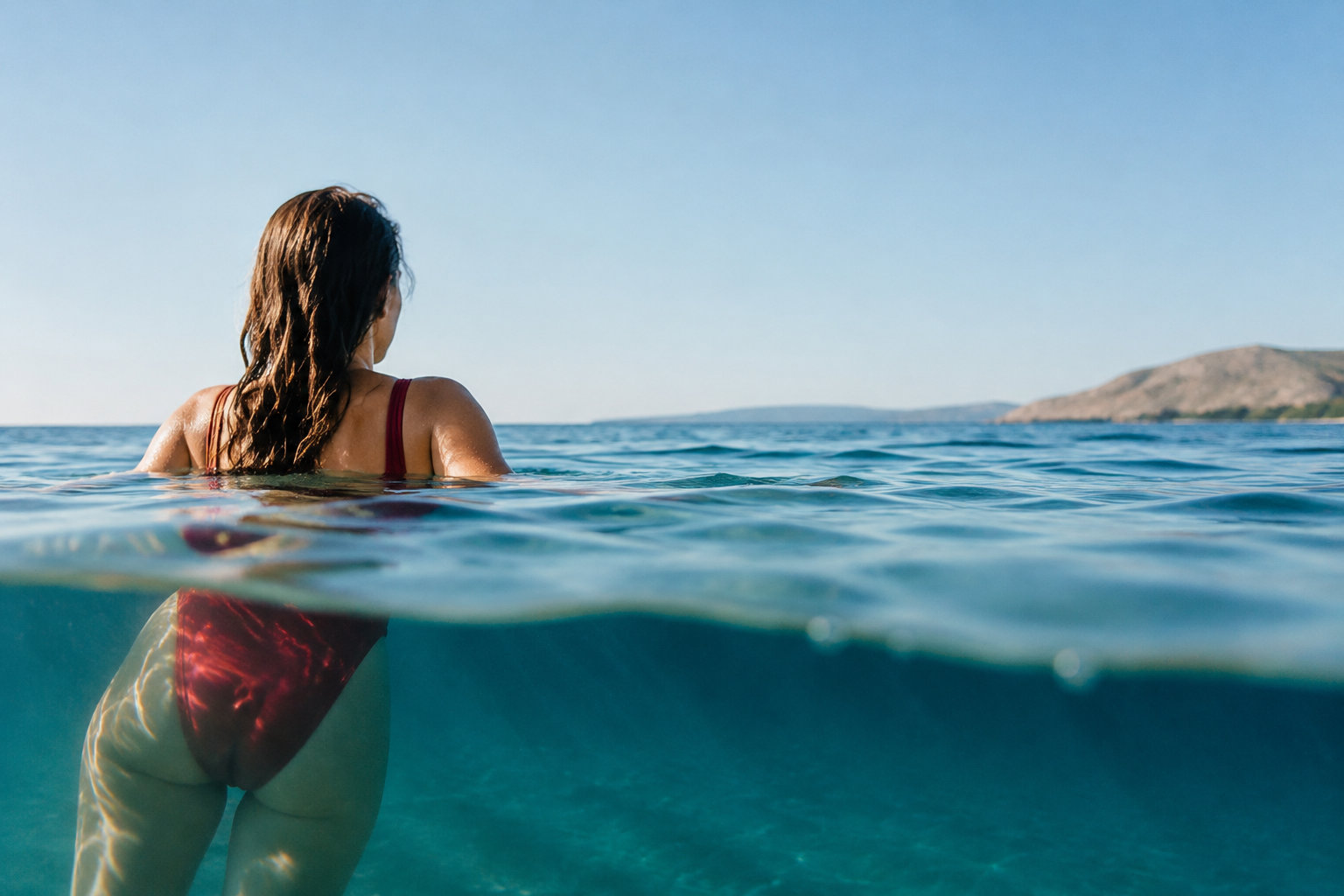 Une femme en maillot de bain rouge vue de dos, à moitié immergée dans une eau turquoise et cristalline.