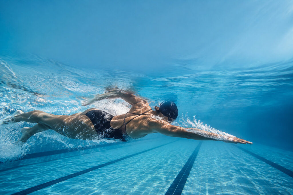 Une nageuse en maillot de bain noir effectue le crawl sous l'eau dans une piscine lumineuse.